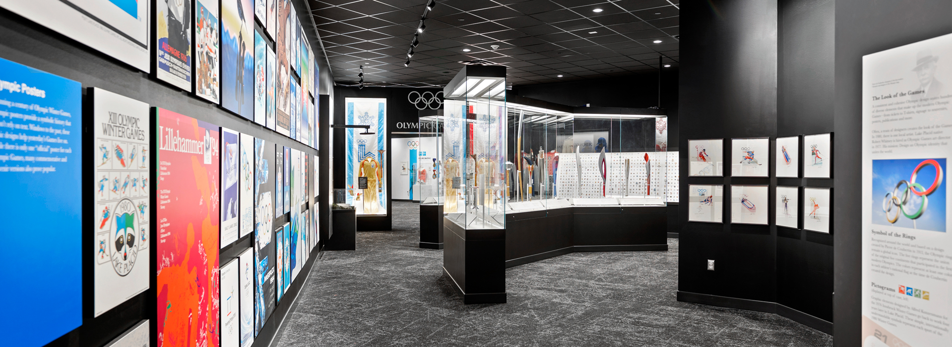 A photograph of the Lake Placid Olympic Museum's Olympic Art Exhibit. The left wall is decorated in a montage of Winter Olympic Posters throughout history. The middle ground has a glass of the Museum's collection of Olympic Torches, and the far right includes water colors from the 1980 Winter Olympics on display. Deep into the background of the image is the archway into The 1980 Games exhibit and a wall of hundreds of Olympic Pins from all over the world.