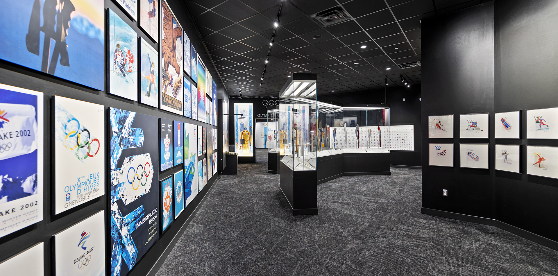 A photograph of the Lake Placid Olympic Museum's Olympic Art Exhibit. The left wall is decorated in a montage of Winter Olympic Posters throughout history. The middle ground has a glass of the Museum's collection of Olympic Torches, and the far right includes water colors from the 1980 Winter Olympics on display. Deep into the background of the image is the archway into The 1980 Games exhibit and a wall of hundreds of Olympic Pins from all over the world.